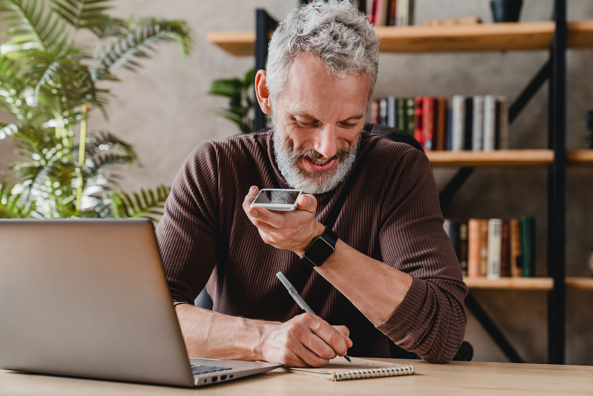 Man writing notes in notepad while talking by speakerphone from workspace